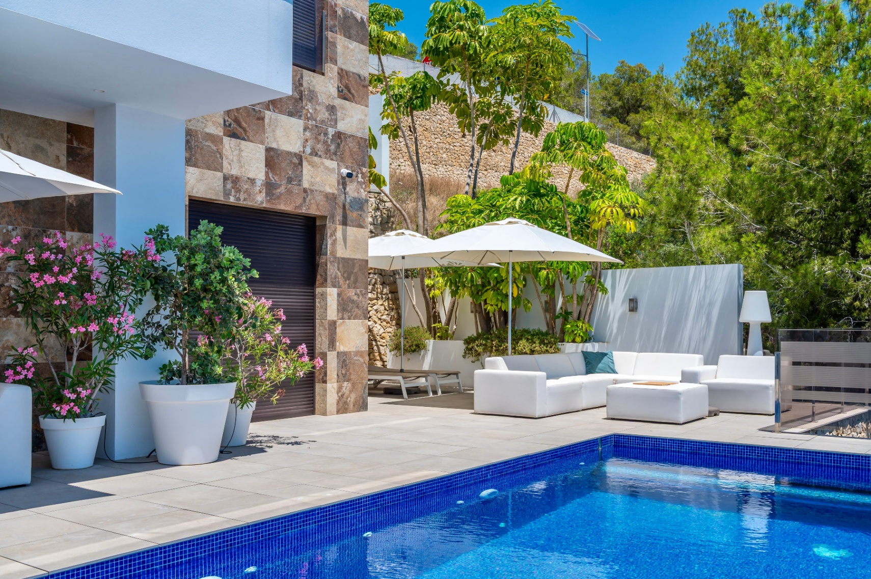 Espaciosa terraza con piscina de mosaico azul y mobiliario exterior blanco bajo sombrillas, complementada por una fachada de piedra y exuberante vegetación mediterránea. Espaciosa terraza con piscina de mosaico azul y mobiliario exterior blanco bajo sombrillas, complementada por una fachada de piedra y exuberante vegetación mediterránea.