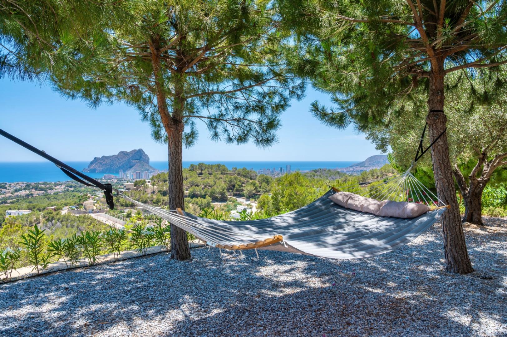 Hamaca acolchada bajo la sombra de pinos, ofreciendo un lugar de descanso con vistas panorámicas al mar Mediterráneo y al icónico Peñon de Ifach. Suelo de grava. Hamaca acolchada bajo la sombra de pinos, ofreciendo un lugar de descanso con vistas panorámicas al mar Mediterráneo y al icónico Peñon de Ifach. Suelo de grava.