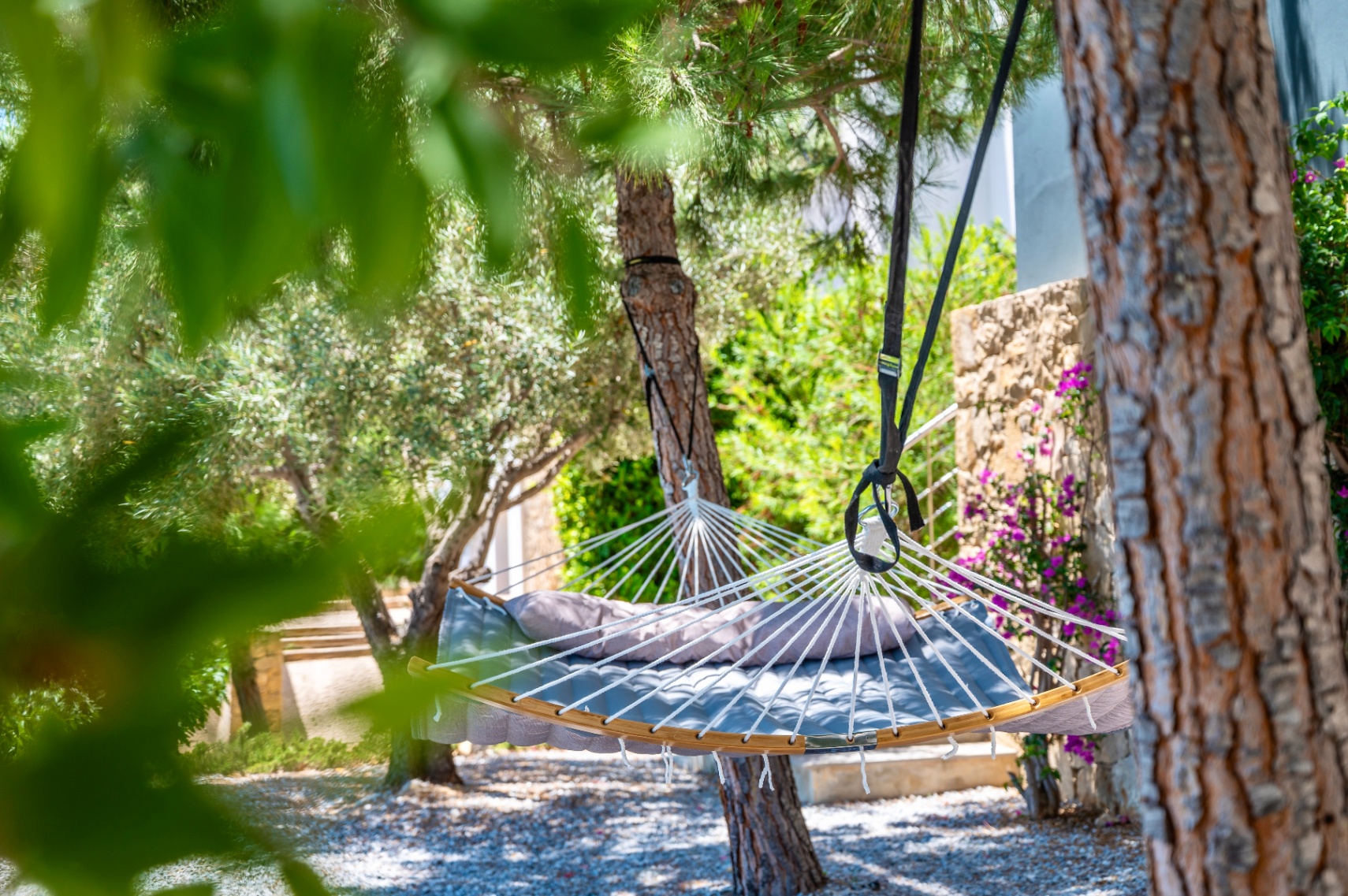 Hamaca acolchada gris con cojín, suspendida entre árboles frondosos sobre suelo de gravilla. Un muro de piedra con flores moradas adorna el fondo del jardín. Hamaca acolchada gris con cojín, suspendida entre árboles frondosos sobre suelo de gravilla. Un muro de piedra con flores moradas adorna el fondo del jardín.