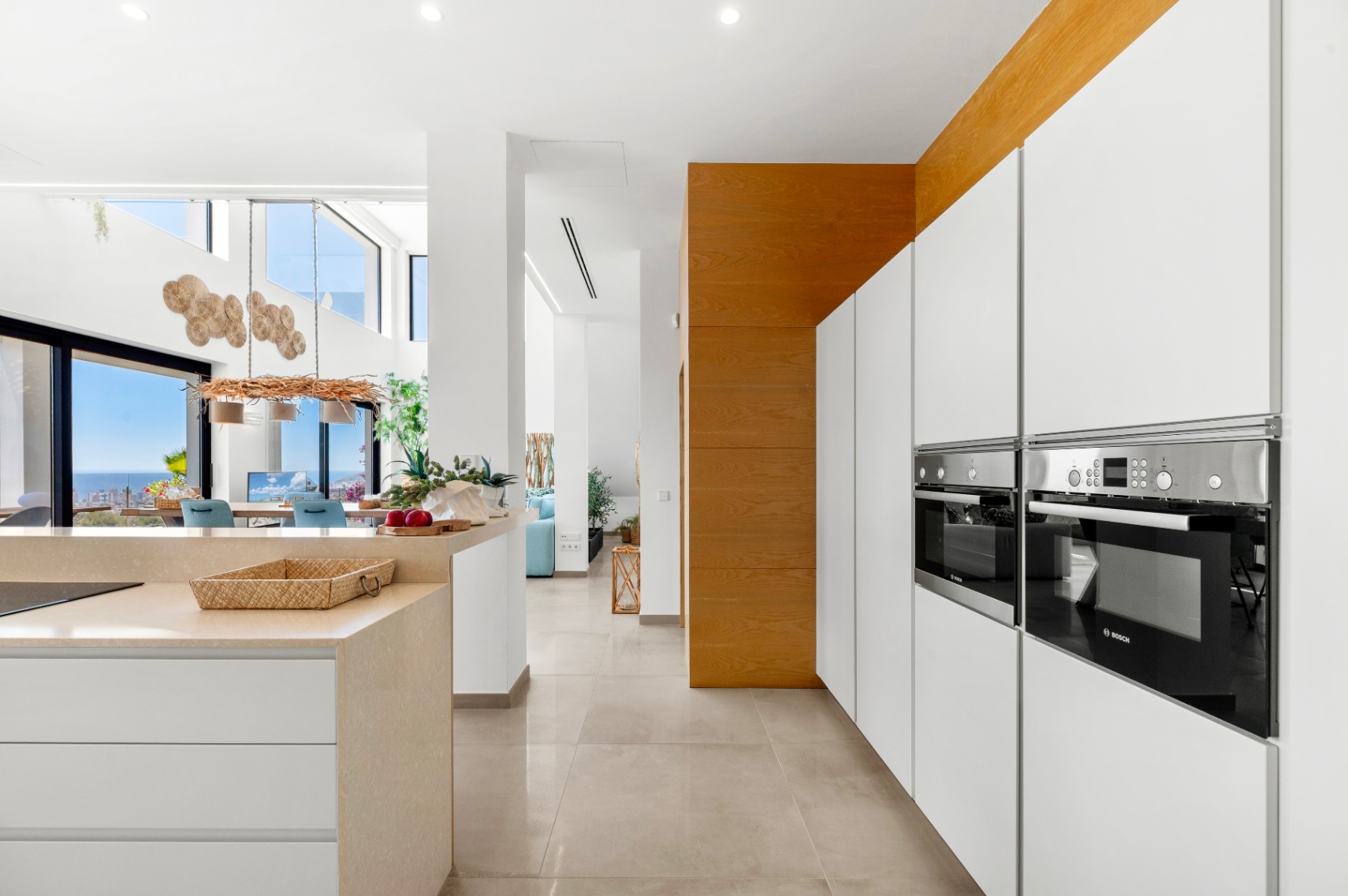 Moderna cocina con elegantes armarios blancos sin tiradores, hornos Bosch integrados y una isla de piedra clara. El diseño abierto conecta con un comedor con vistas panorámicas al mar. Moderna cocina con elegantes armarios blancos sin tiradores, hornos Bosch integrados y una isla de piedra clara. El diseño abierto conecta con un comedor con vistas panorámicas al mar.