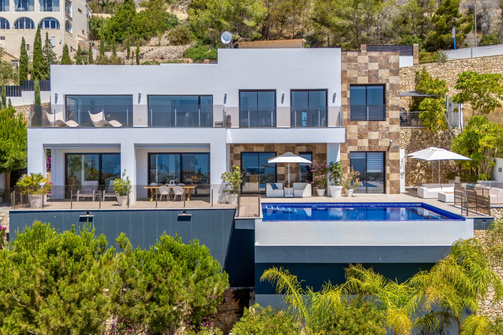Moderna villa de varios niveles con fachada blanca y detalles de piedra natural. Dispone de piscina infinita, solárium con mobiliario blanco y dos terrazas acristaladas para comedor exterior y relax entre vegetación. Moderna villa de varios niveles con fachada blanca y detalles de piedra natural. Dispone de piscina infinita, solárium con mobiliario blanco y dos terrazas acristaladas para comedor exterior y relax entre vegetación.