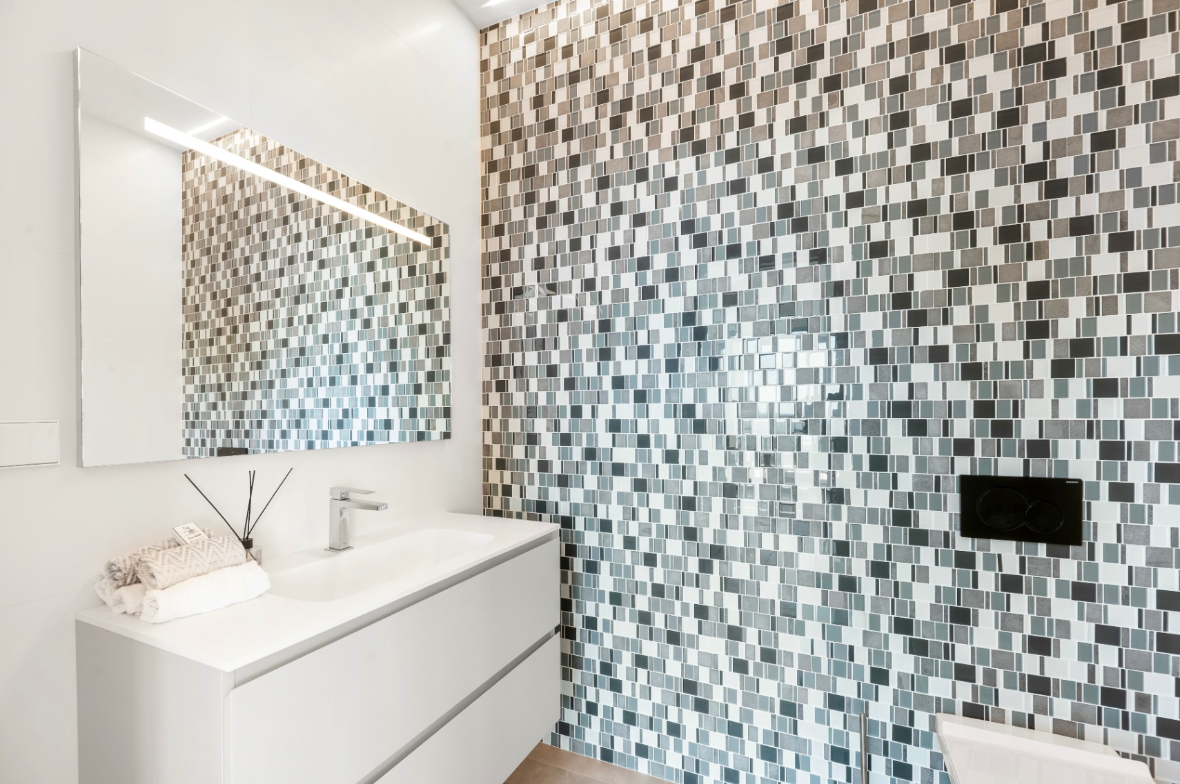 Moderno cuarto de baño con mueble flotante blanco, lavabo integrado y grifo cromado. La pared destaca por su revestimiento de mosaico en tonos grises y blancos, con espejo iluminado. Moderno cuarto de baño con mueble flotante blanco, lavabo integrado y grifo cromado. La pared destaca por su revestimiento de mosaico en tonos grises y blancos, con espejo iluminado.