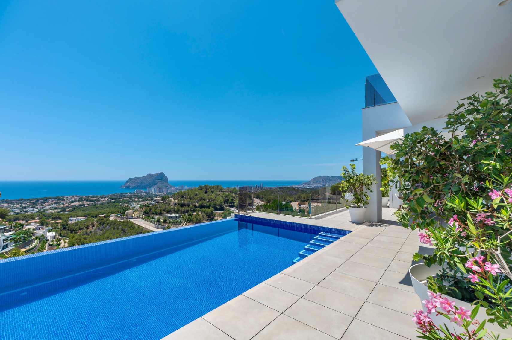 Piscina infinita con mosaico azul y amplia terraza de baldosas claras. Ofrece vistas panorámicas despejadas al mar y al Peñón de Ifach, con arquitectura moderna y flora mediterránea. Piscina infinita con mosaico azul y amplia terraza de baldosas claras. Ofrece vistas panorámicas despejadas al mar y al Peñón de Ifach, con arquitectura moderna y flora mediterránea.