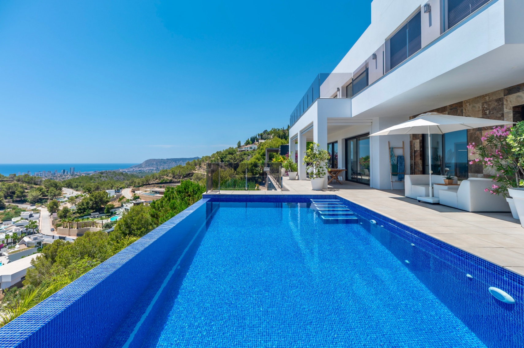 Piscina infinita de mosaico azul con escalones integrados, flanqueada por una amplia terraza con mobiliario lounge y vistas panorámicas a la costa y la ciudad. Piscina infinita de mosaico azul con escalones integrados, flanqueada por una amplia terraza con mobiliario lounge y vistas panorámicas a la costa y la ciudad.