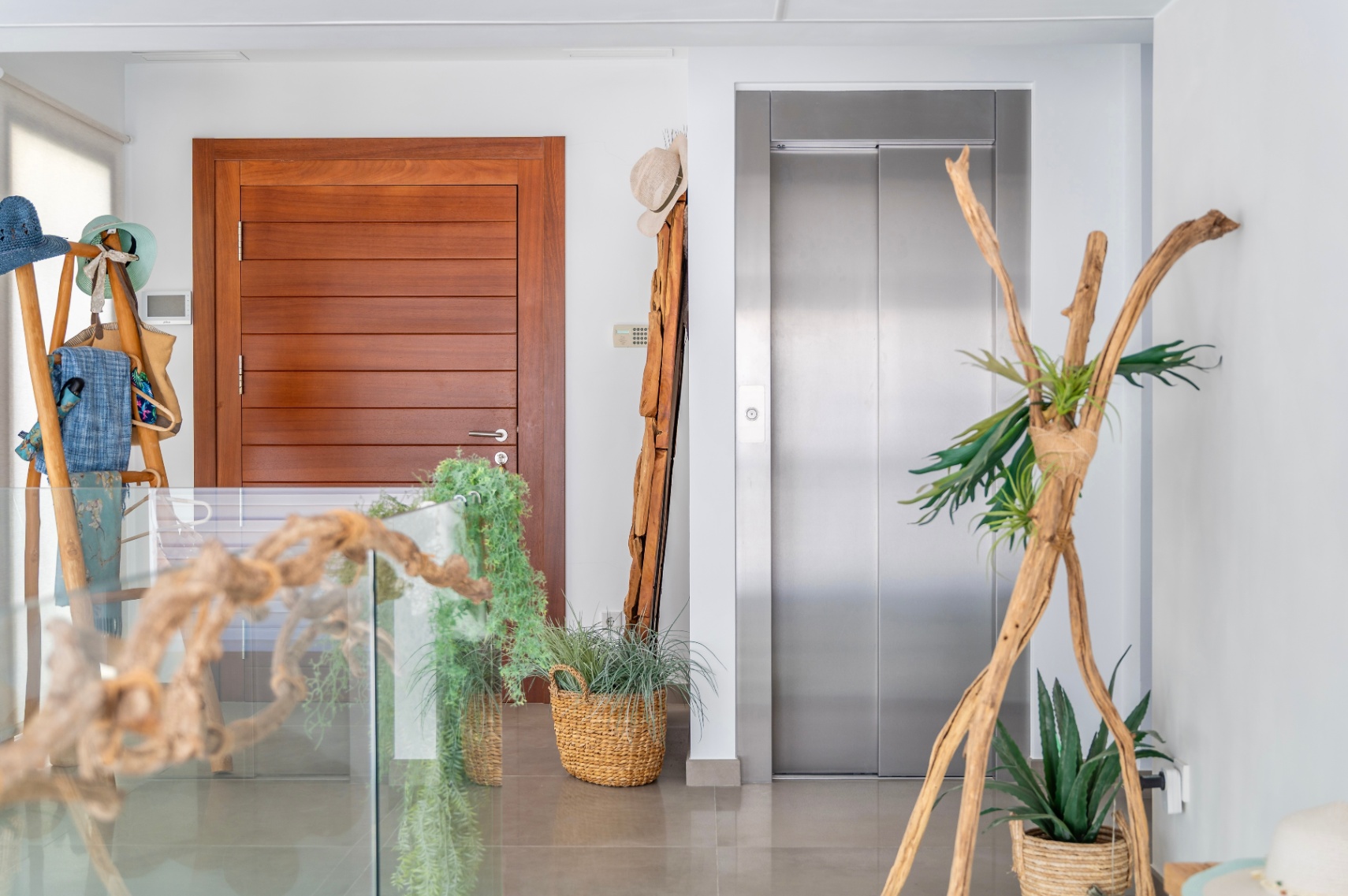 Recibidor interior moderno con puerta de madera lisa y ascensor de acero inoxidable. El espacio se decora elegantemente con elementos naturales como maderas flotantes, plantas en cestas y una barandilla de cristal. Recibidor interior moderno con puerta de madera lisa y ascensor de acero inoxidable. El espacio se decora elegantemente con elementos naturales como maderas flotantes, plantas en cestas y una barandilla de cristal.