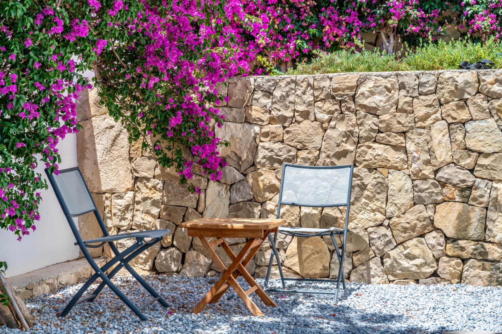 Rincón exterior con dos sillas plegables grises y mesa de madera sobre grava. Un muro de piedra natural sirve de fondo a vibrantes buganvillas moradas. Rincón exterior con dos sillas plegables grises y mesa de madera sobre grava. Un muro de piedra natural sirve de fondo a vibrantes buganvillas moradas.