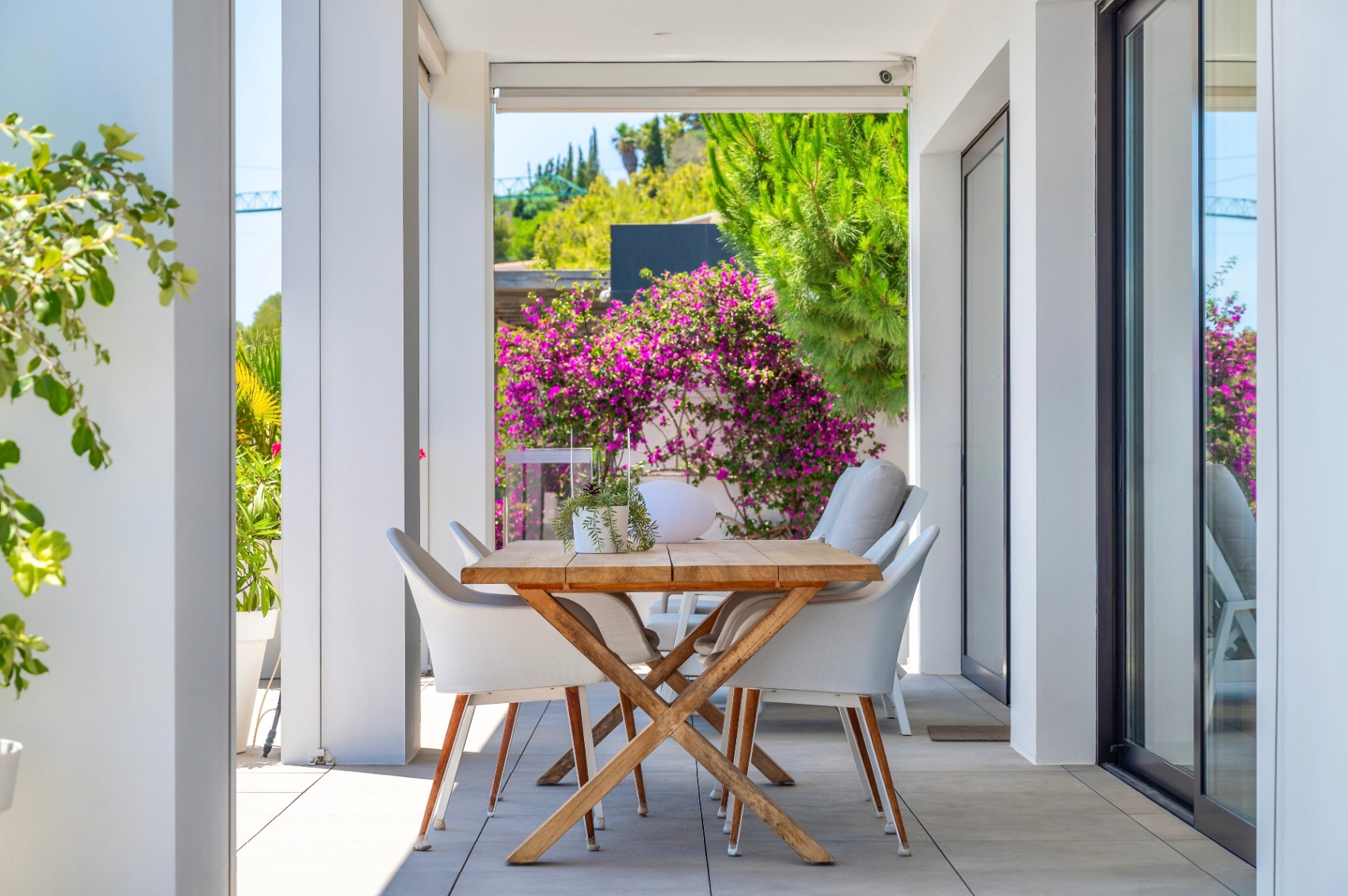 Terraza cubierta con mesa de comedor de madera y sillas blancas, enmarcada por exuberante vegetación y vibrantes buganvillas, ideal para comidas al aire libre. Terraza cubierta con mesa de comedor de madera y sillas blancas, enmarcada por exuberante vegetación y vibrantes buganvillas, ideal para comidas al aire libre.