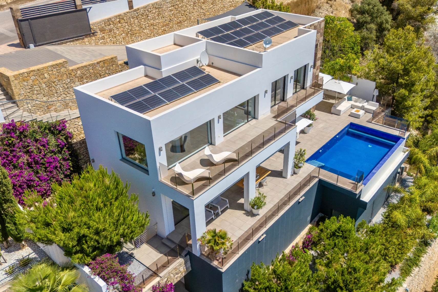 Vista aérea de una villa moderna blanca con múltiples niveles aterrazados, piscina rectangular y amplias zonas de estar exteriores. Paneles solares en los tejados planos, rodeada de exuberante paisajismo mediterráneo. Vista aérea de una villa moderna blanca con múltiples niveles aterrazados, piscina rectangular y amplias zonas de estar exteriores. Paneles solares en los tejados planos, rodeada de exuberante paisajismo mediterráneo.