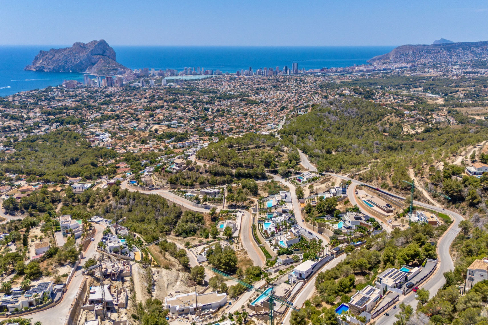 Vista aérea panorámica de Calpe con el Peñón de Ifach, la costa y una zona residencial con villas y piscinas entre colinas verdes y carreteras serpenteantes. Vista aérea panorámica de Calpe con el Peñón de Ifach, la costa y una zona residencial con villas y piscinas entre colinas verdes y carreteras serpenteantes.
