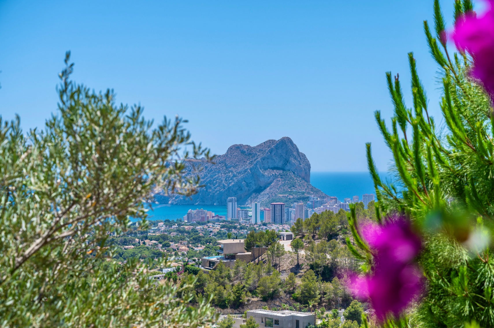 Vista panorámica del Peñón de Ifach y la costa de Calpe, enmarcada por vegetación mediterránea y con villas modernas en la ladera. Vista panorámica del Peñón de Ifach y la costa de Calpe, enmarcada por vegetación mediterránea y con villas modernas en la ladera.