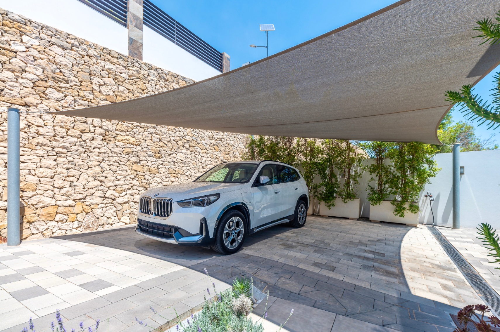 Zona de aparcamiento exterior con una vela de sombra y pavimento de adoquines bitono. Un muro de piedra natural y jardineras con vegetación delimitan el espacio. Zona de aparcamiento exterior con una vela de sombra y pavimento de adoquines bitono. Un muro de piedra natural y jardineras con vegetación delimitan el espacio.