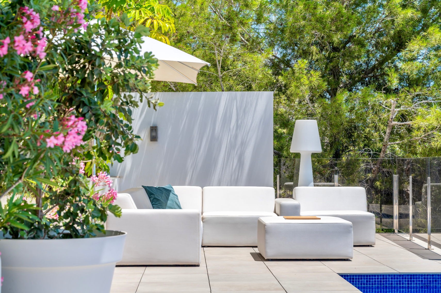 Zona lounge exterior con sofás modulares blancos y otomana en patio de baldosas. Rodeado de vegetación y una planta con flores rosas, junto a una piscina y barandilla de cristal. Zona lounge exterior con sofás modulares blancos y otomana en patio de baldosas. Rodeado de vegetación y una planta con flores rosas, junto a una piscina y barandilla de cristal.
