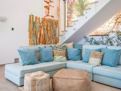 Acogedora sala de estar con sofá seccional azul claro, cojines variados y dos pufs de cuero. Escalera moderna de cristal junto a una pantalla de madera y plantas, con muro de piedra visible.