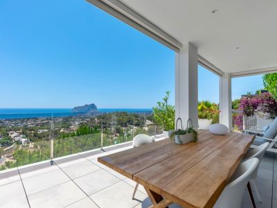 Amplia terraza cubierta con mesa de comedor de madera maciza y sillas blancas. Ofrece vistas panorámicas al mar, Calpe y el Peñón de Ifach a través de barandilla de cristal.