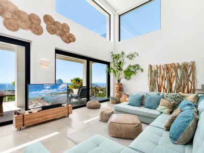 Amplio salón con techos altos, grandes ventanales y puertas correderas que ofrecen vistas panorámicas al mar. Un sofá seccional azul claro se combina con detalles de madera y arte mural.