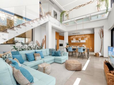 Amplio salón de planta abierta con sofá seccional azul claro, escalera moderna de cristal y cocina con detalles de madera cálida, todo iluminado por luz natural.