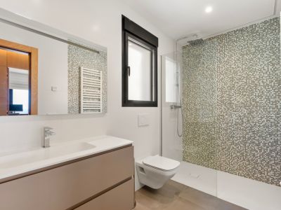 Baño moderno con ducha a ras de suelo y pared de mosaico en tonos tierra. Incluye un mueble de lavabo flotante con lavamanos integrado, inodoro suspendido y ventana con marco negro.
