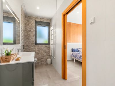 Baño moderno con lavabo gris, espejo iluminado y azulejos texturizados. Incluye una ventana esmerilada, toallero calefactado y acceso directo a un dormitorio.