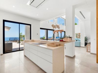 Cocina de diseño abierta con isla central de encimera beige y placa de inducción. Grandes ventanales correderos ofrecen vistas directas al mar Mediterráneo desde un espacio luminoso y moderno.