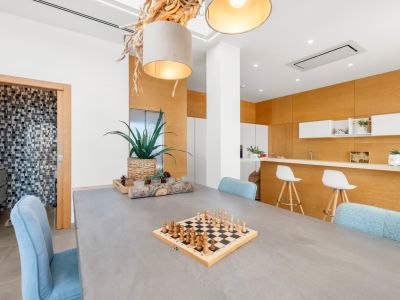 Comedor moderno con mesa de hormigón y sillas azules. Destaca una lámpara colgante de madera natural y una cocina abierta con panelado de madera clara y barra. Al fondo, un baño con mosaicos.