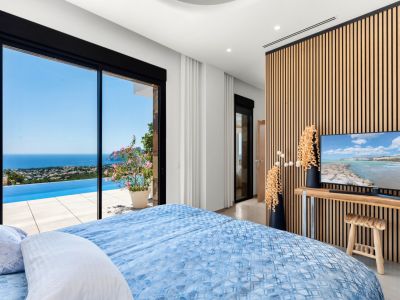 Dormitorio con amplias puertas correderas de cristal que se abren a una piscina infinita y vistas panorámicas al mar. Una moderna pared de lamas de madera enmarca la televisión, complementando el interior luminoso.