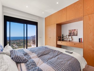 Dormitorio principal con amplias puertas correderas de cristal y vistas panorámicas al mar y la costa. Armarios empotrados de madera clara con escritorio integrado.