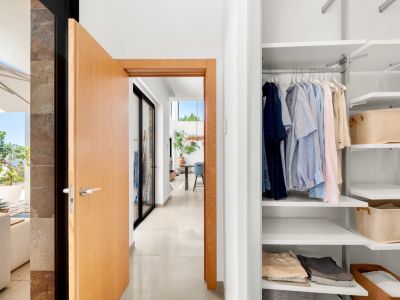 Entrada a un moderno pasillo con puerta de madera clara abierta, mostrando un armario empotrado blanco con estantes ajustables y una terraza exterior soleada con vistas al mar.