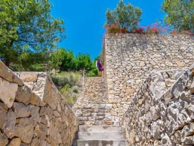 Escalera exterior de piedra natural flanqueada por muros de mampostería seca, ascendiendo entre exuberante vegetación mediterránea bajo un cielo azul claro.