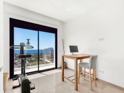 Espacio multifuncional con escritorio alto de madera y bicicleta estática, ambos orientados a las amplias vistas al mar y al Peñon de Ifach a través de las grandes puertas correderas de cristal.