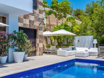 Espaciosa terraza con piscina de mosaico azul y mobiliario exterior blanco bajo sombrillas, complementada por una fachada de piedra y exuberante vegetación mediterránea.