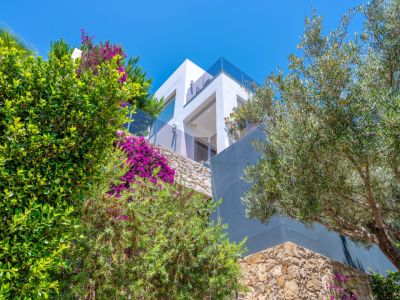 Exterior de villa moderna con fachadas blancas y grises, y barandillas de cristal en terrazas elevadas. El exuberante paisajismo mediterráneo, con buganvillas y un olivo, se integra con el muro de piedra.