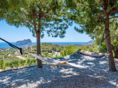 Hamaca acolchada bajo la sombra de pinos, ofreciendo un lugar de descanso con vistas panorámicas al mar Mediterráneo y al icónico Peñon de Ifach. Suelo de grava.