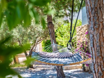 Hamaca acolchada gris con cojín, suspendida entre árboles frondosos sobre suelo de gravilla. Un muro de piedra con flores moradas adorna el fondo del jardín.