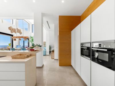 Moderna cocina con elegantes armarios blancos sin tiradores, hornos Bosch integrados y una isla de piedra clara. El diseño abierto conecta con un comedor con vistas panorámicas al mar.