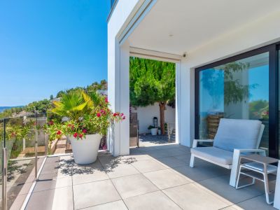 Moderna terraza exterior con suelo de baldosas grises claras y barandilla de cristal, ofreciendo vistas despejadas a la ladera verde. Incluye asientos cómodos y plantas en maceta bajo luz natural.
