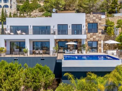 Moderna villa de varios niveles con fachada blanca y detalles de piedra natural. Dispone de piscina infinita, solárium con mobiliario blanco y dos terrazas acristaladas para comedor exterior y relax entre vegetación.