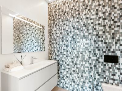 Moderno cuarto de baño con mueble flotante blanco, lavabo integrado y grifo cromado. La pared destaca por su revestimiento de mosaico en tonos grises y blancos, con espejo iluminado.