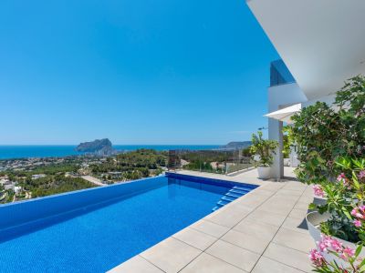 Piscina infinita con mosaico azul y amplia terraza de baldosas claras. Ofrece vistas panorámicas despejadas al mar y al Peñón de Ifach, con arquitectura moderna y flora mediterránea.
