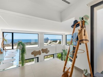 Planta superior con suelo de gres claro y barandilla de cristal, ofreciendo vistas panorámicas al mar y al Peñon de Ifach. Incluye una pasarela de cristal con peldaños ovalados y un perchero de madera.