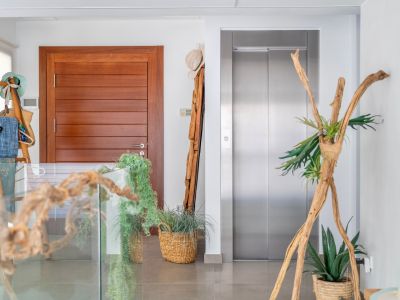Recibidor interior moderno con puerta de madera lisa y ascensor de acero inoxidable. El espacio se decora elegantemente con elementos naturales como maderas flotantes, plantas en cestas y una barandilla de cristal.