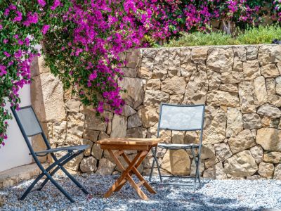 Rincón exterior con dos sillas plegables grises y mesa de madera sobre grava. Un muro de piedra natural sirve de fondo a vibrantes buganvillas moradas.