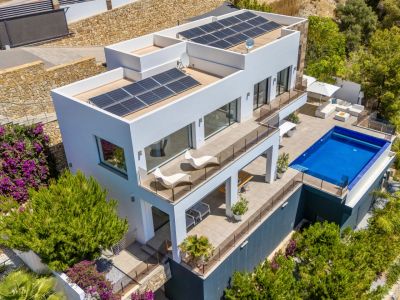Vista aérea de una villa moderna blanca con múltiples niveles aterrazados, piscina rectangular y amplias zonas de estar exteriores. Paneles solares en los tejados planos, rodeada de exuberante paisajismo mediterráneo.