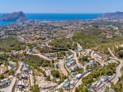 Vista aérea panorámica de Calpe con el Peñón de Ifach, la costa y una zona residencial con villas y piscinas entre colinas verdes y carreteras serpenteantes.