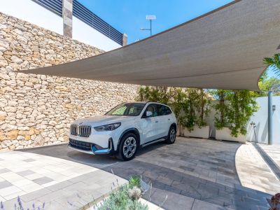 Zona de aparcamiento exterior con una vela de sombra y pavimento de adoquines bitono. Un muro de piedra natural y jardineras con vegetación delimitan el espacio.