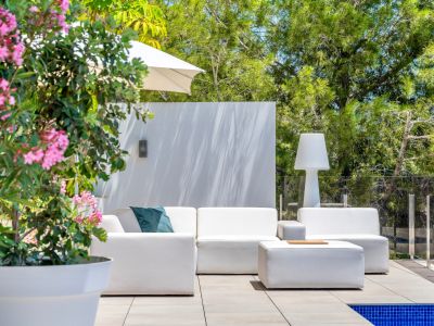 Zona lounge exterior con sofás modulares blancos y otomana en patio de baldosas. Rodeado de vegetación y una planta con flores rosas, junto a una piscina y barandilla de cristal.
