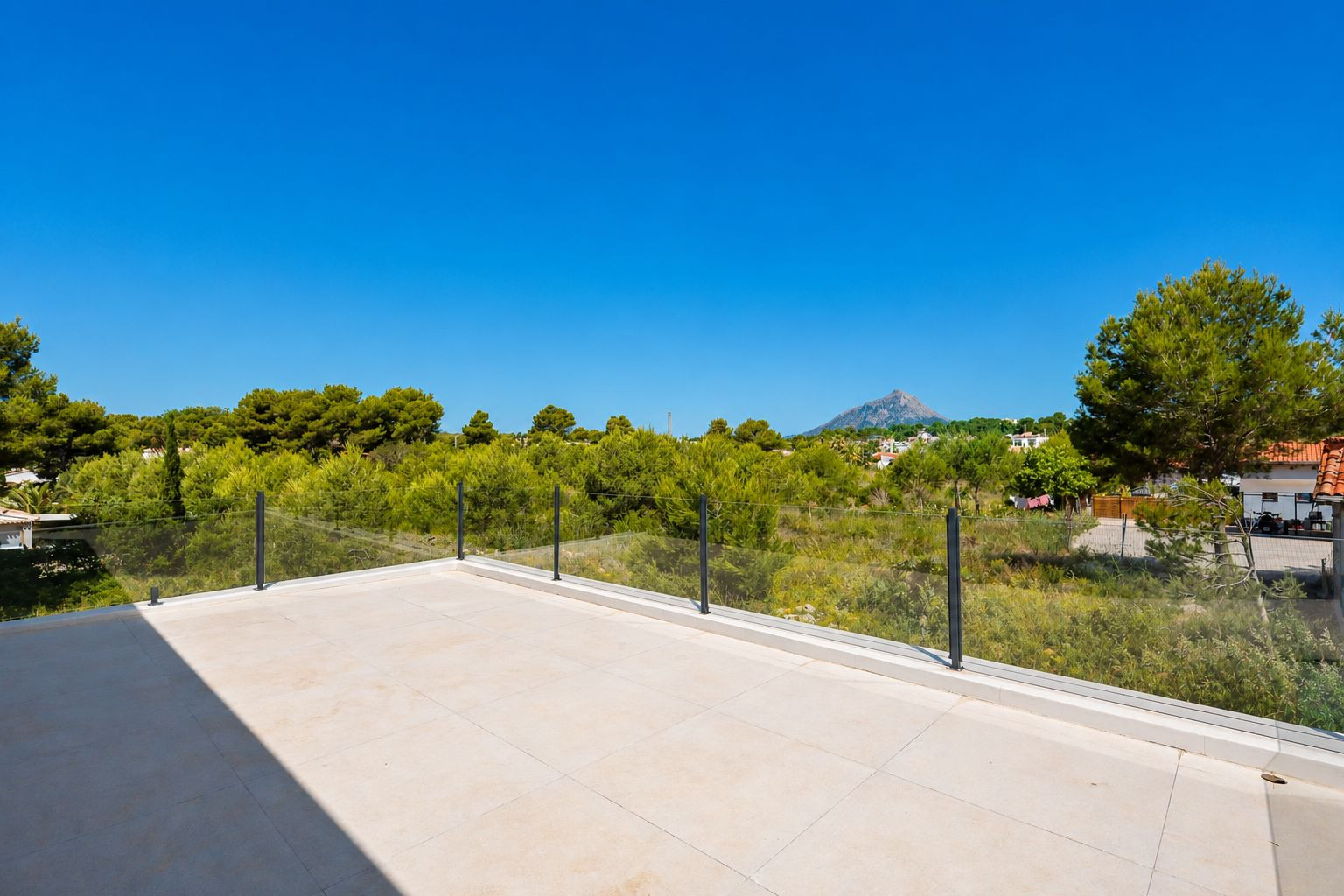 Amplia terraza superior con suelo de gres porcelánico claro y barandilla de cristal, ofreciendo vistas a la vegetación mediterránea y al perfil de la montaña.