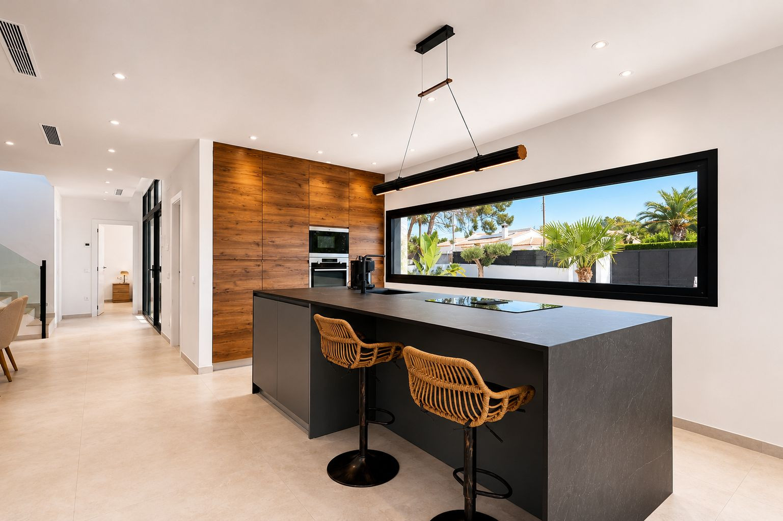 Cocina moderna con isla central de encimera oscura y taburetes de ratán. Pared de madera con electrodomésticos integrados y ventanal horizontal con vistas al exterior.