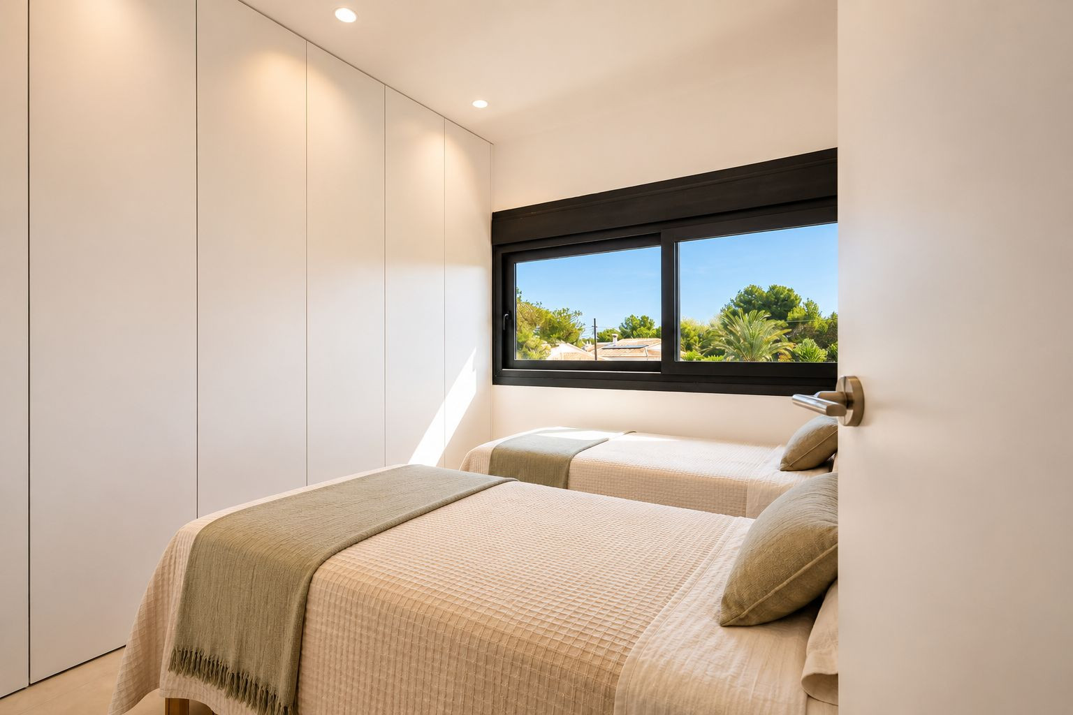 Dormitorio secundario con dos camas individuales, armarios empotrados blancos sin tiradores y ventana con vistas a la vegetación mediterránea. Iluminación empotrada y luz natural.