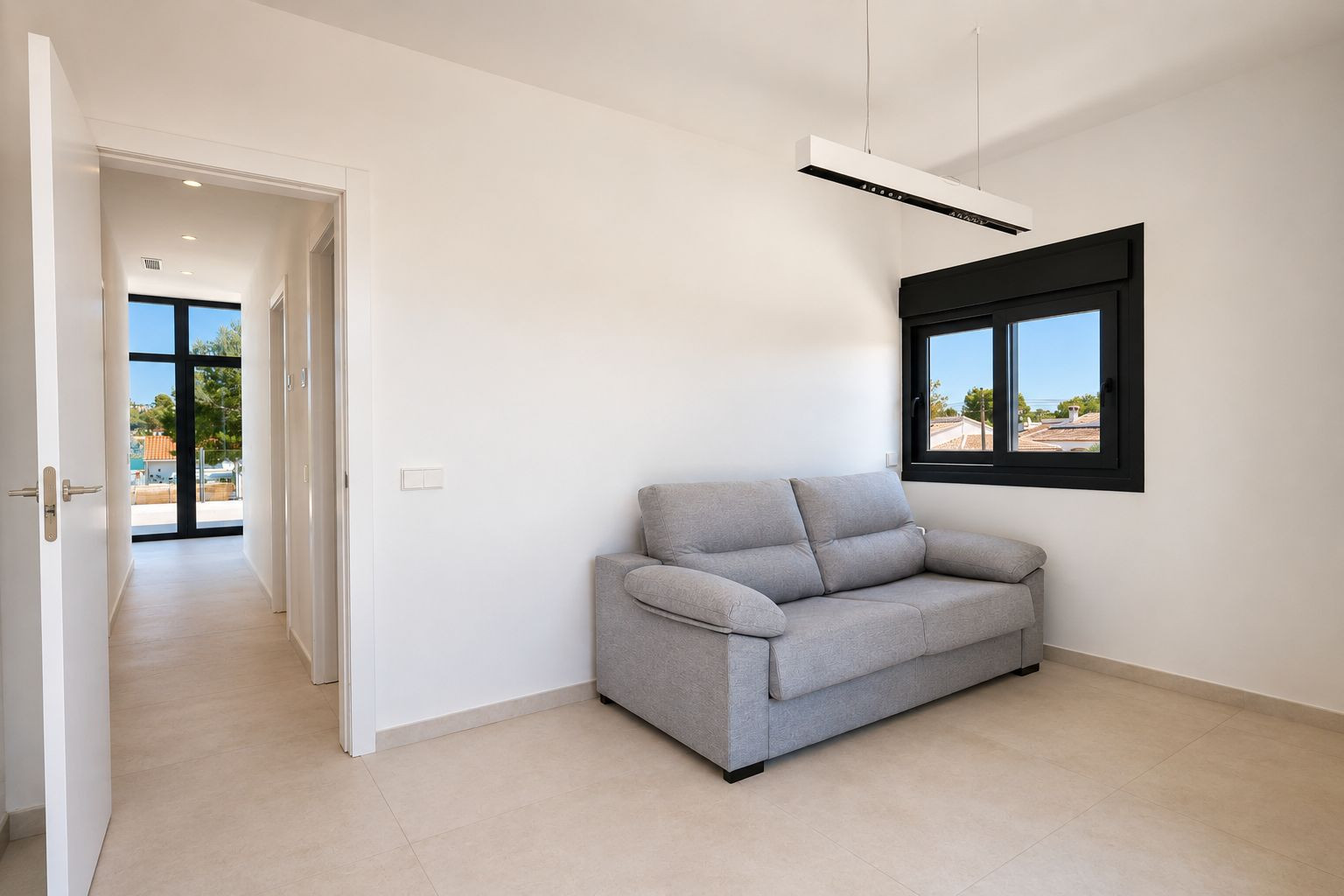 Espacio auxiliar luminoso con sofá gris, ventana de marco negro y vistas al entorno. El pasillo adyacente se abre a una gran ventana exterior, destacando la luz natural.