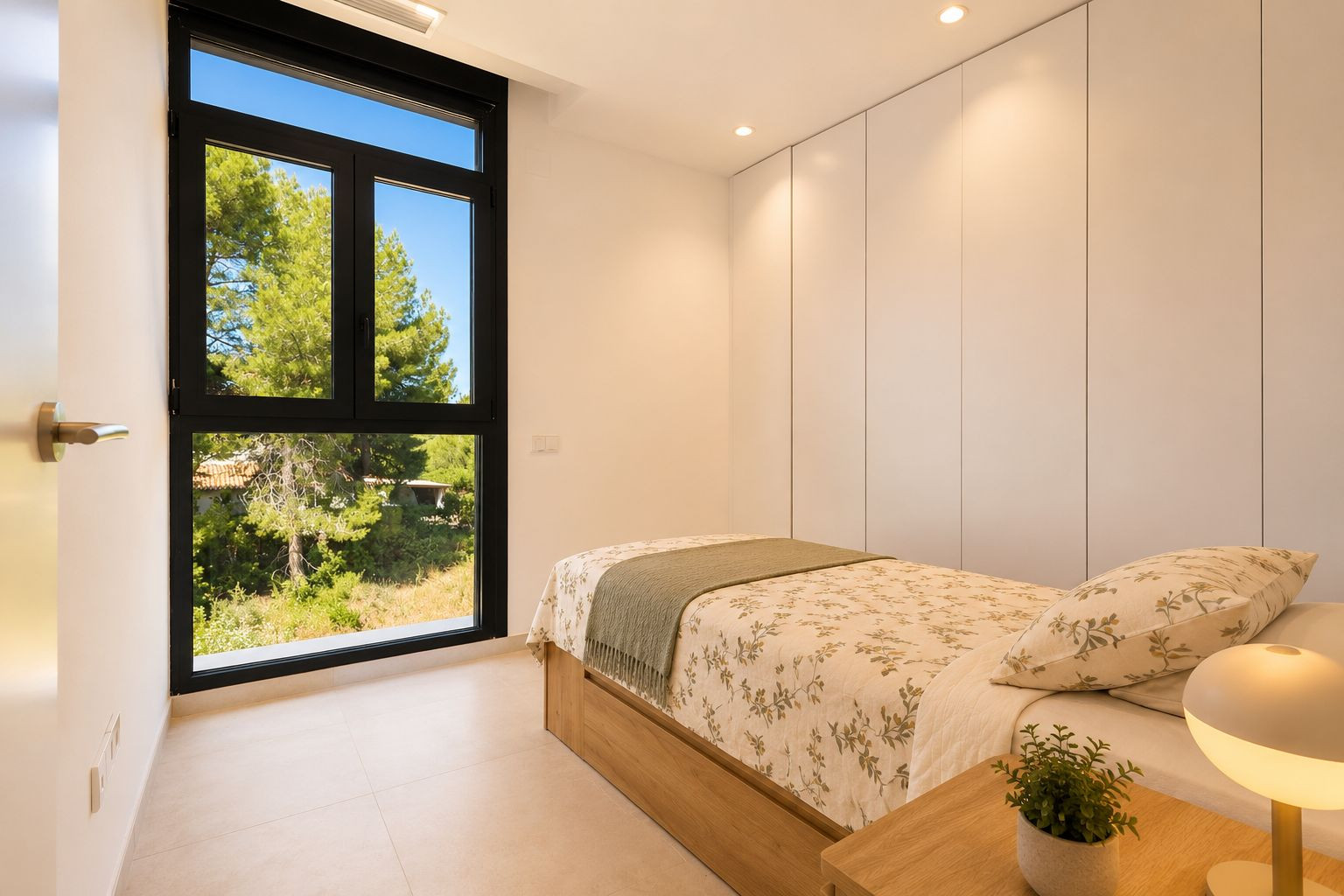Luminoso dormitorio con cama individual de base de madera, armarios empotrados blancos y gran ventanal negro con vistas a la naturaleza. Iluminación empotrada en el techo.